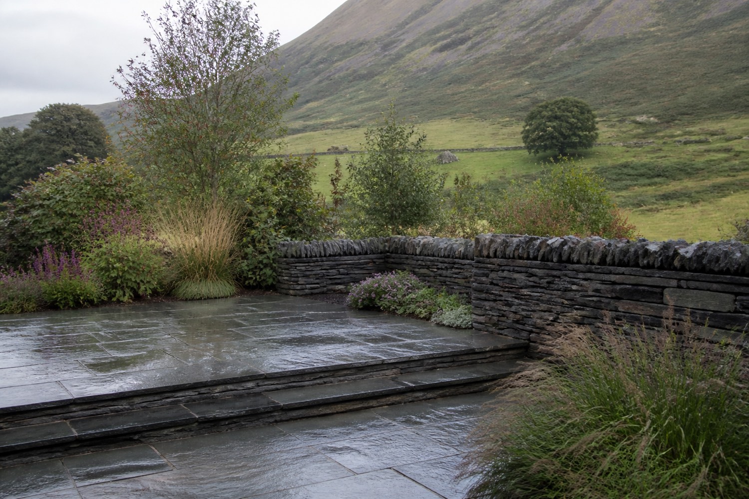 A two-level riven slate terrace with a re-coursed dry stone wall, planting and a fell rising behind